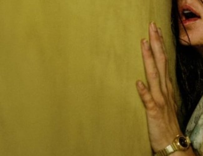 Image of a blank yellow wall, with a woman pressed up against it, looking distraught, and only half of her face and body shown.