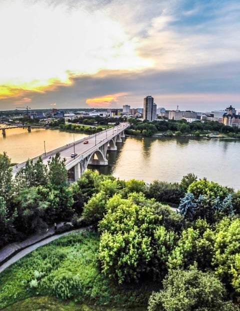 Aerial shot of Saskatoon with a green park, bridge, and water in the foreground