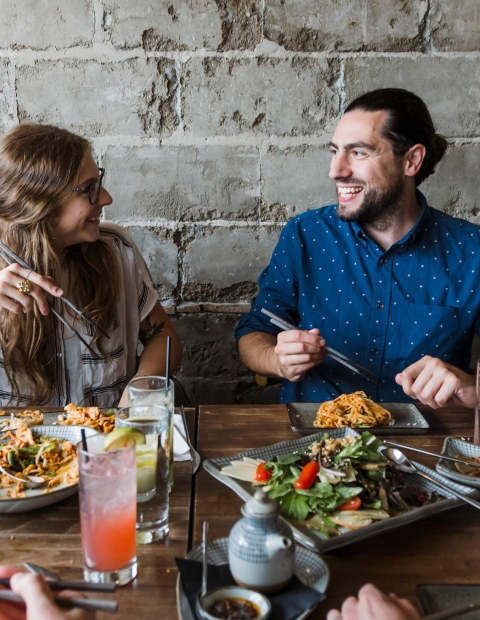 Couple sitting in front of a grey brick wall, at a wooden dining table eating