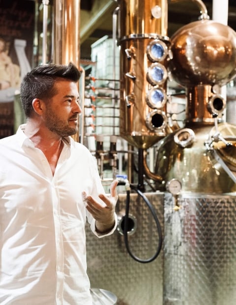 a man in a white shirt standing in front of a beer distiller, giving a tour to guests. 