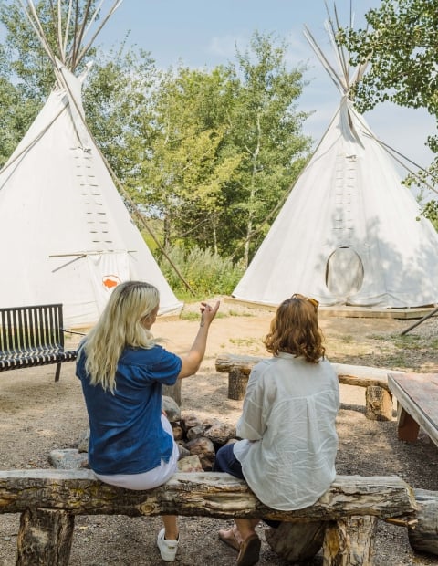 two women sitting on a bench looking at a teepee