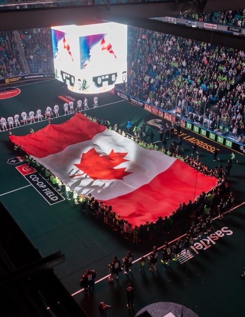 A football stadium fulled with fans. There is a demonstration on the field were individuals are holding a huge Canadian flag