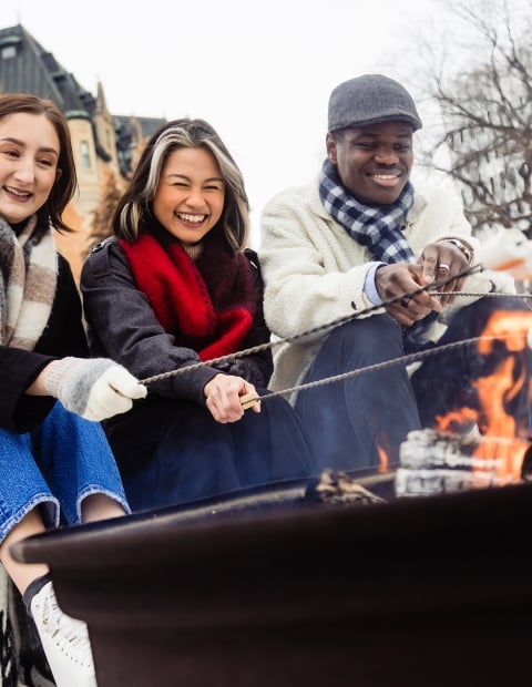 Three individuals sitting around an outdoor fire pit roasting marshmallows in the winter