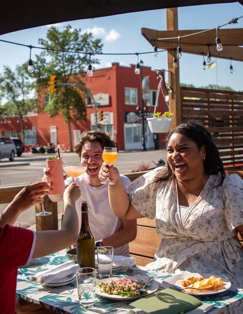four individuals sitting at an outdoor table on a restaurant table "cheersing" drinks