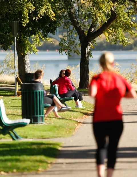 Two women running in a park. In the background some additional individuals are sitting on a park bench.