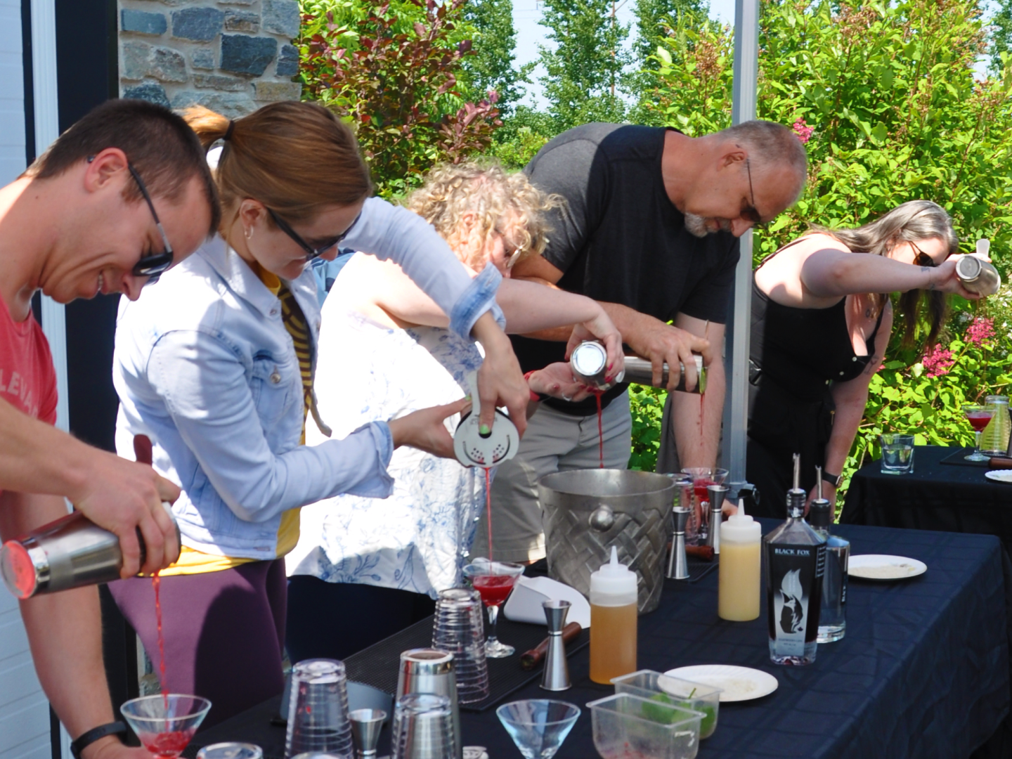 Five individuals outside pouring cocktails into a glass that they made during a Cocktail Class