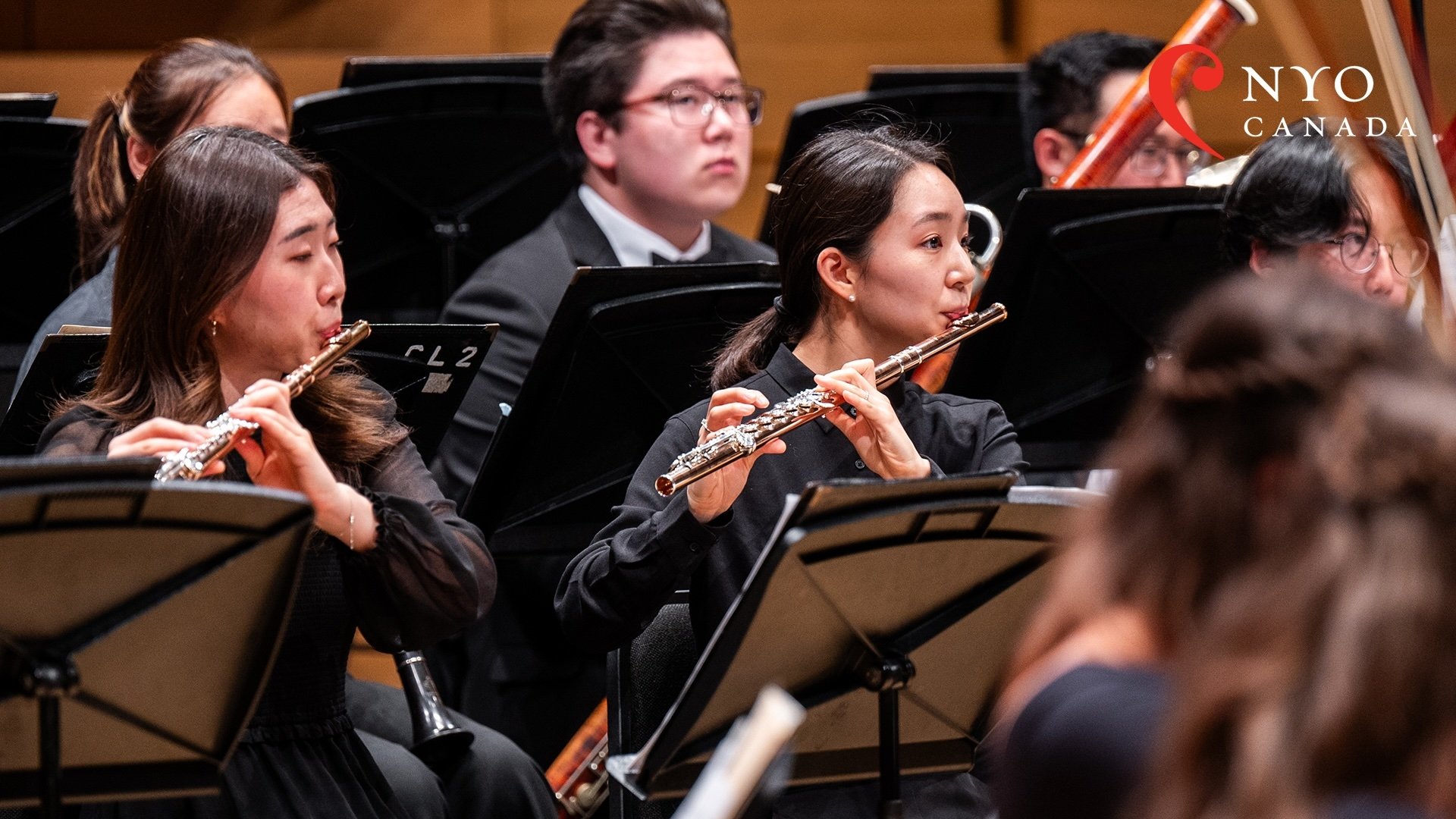 Two flutists in focus, playing their instruments onstage, surrounded by the rest of the woodwind section in performance.