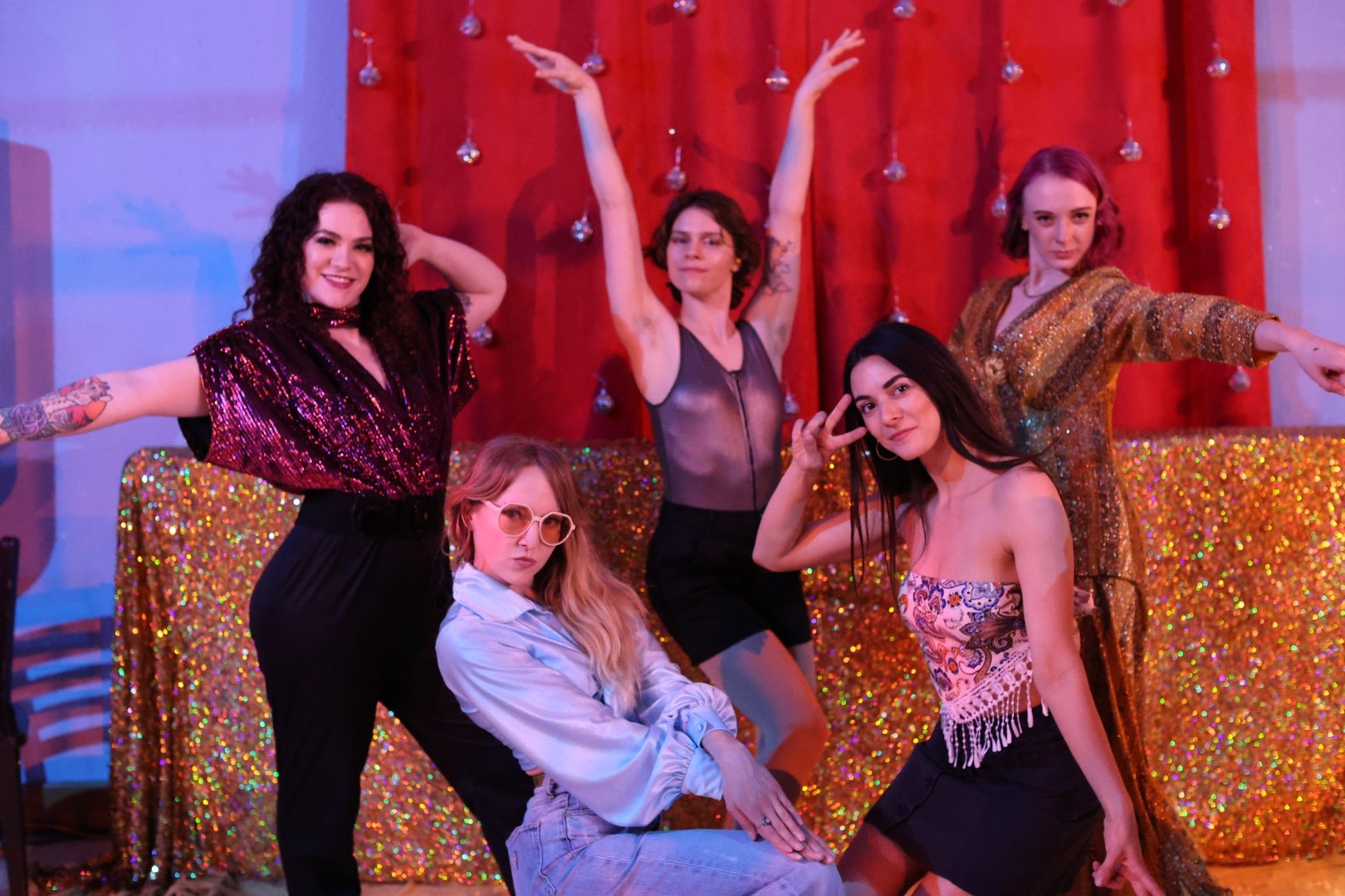 5 dancers wear 70's inspired outfits and strike disco dance poses in front of a gold glittery backdrop.