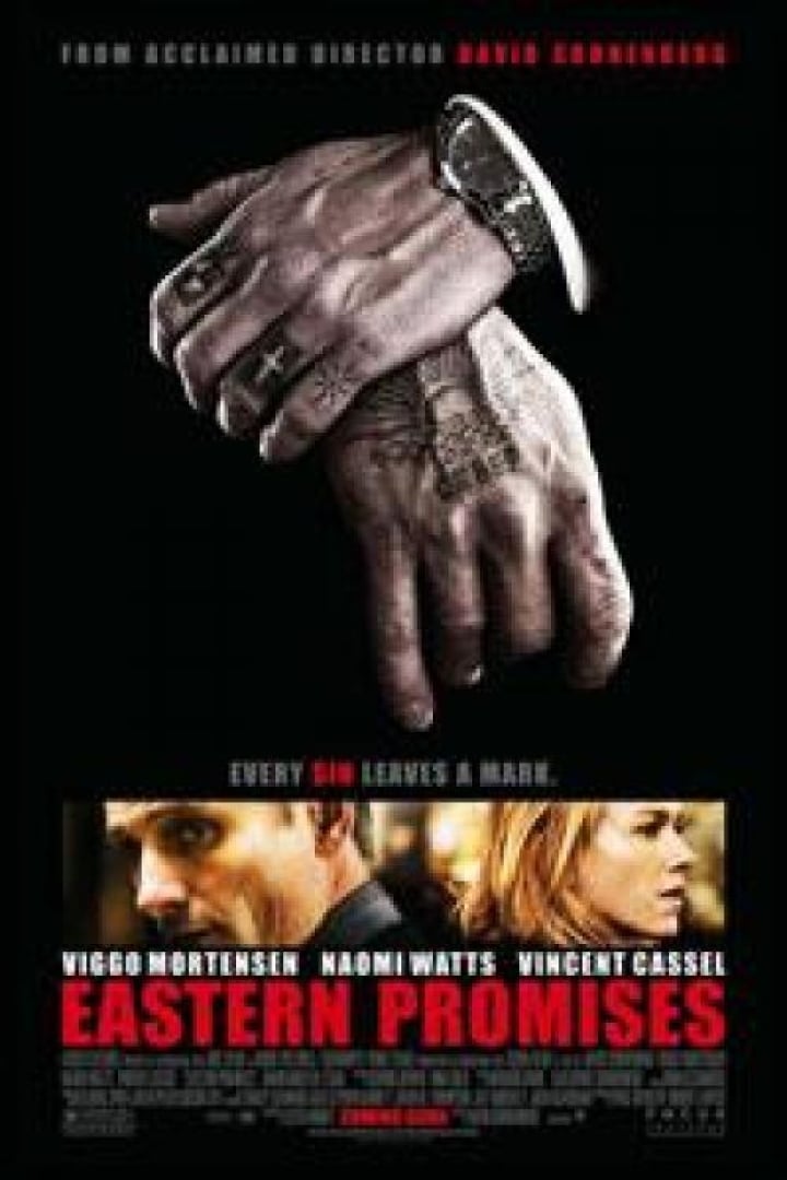 Image from the movie eastern Promises with a man holding his wrist, wearing  a black suit and an expensive watch. His hands are covered in tattoos. 