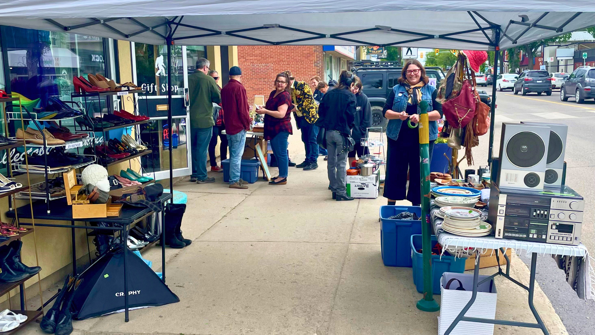 picture of people smiling and talking on the sidewalk with a market tent, shelves, and tables with goods to shop