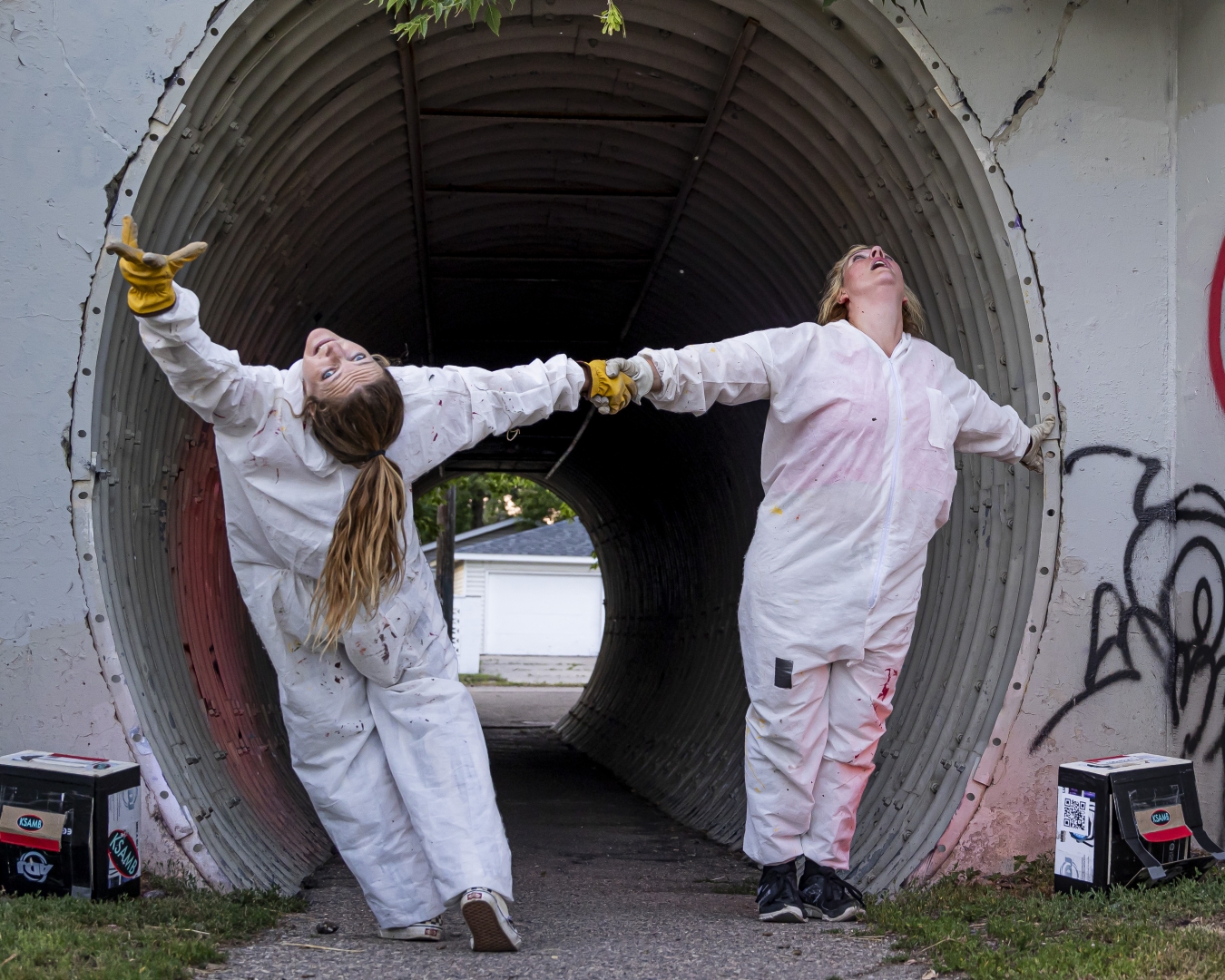 2 dancers in white paint suits hold hands and bend backwards in a metal underpass tunnel