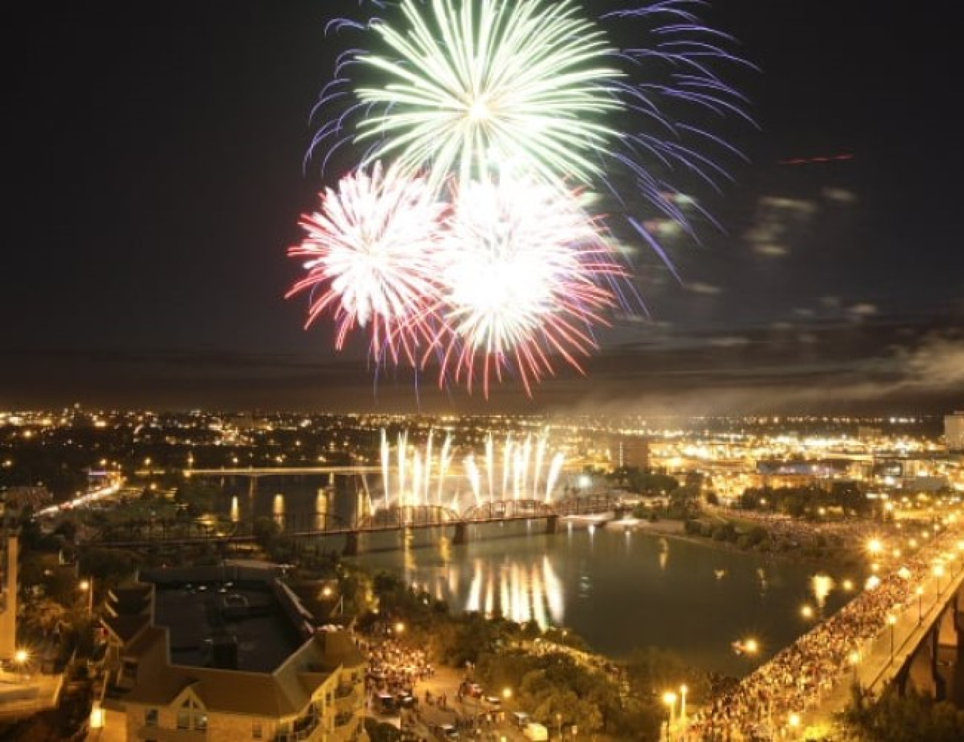 Large crowd watching fireworks over a river at night.