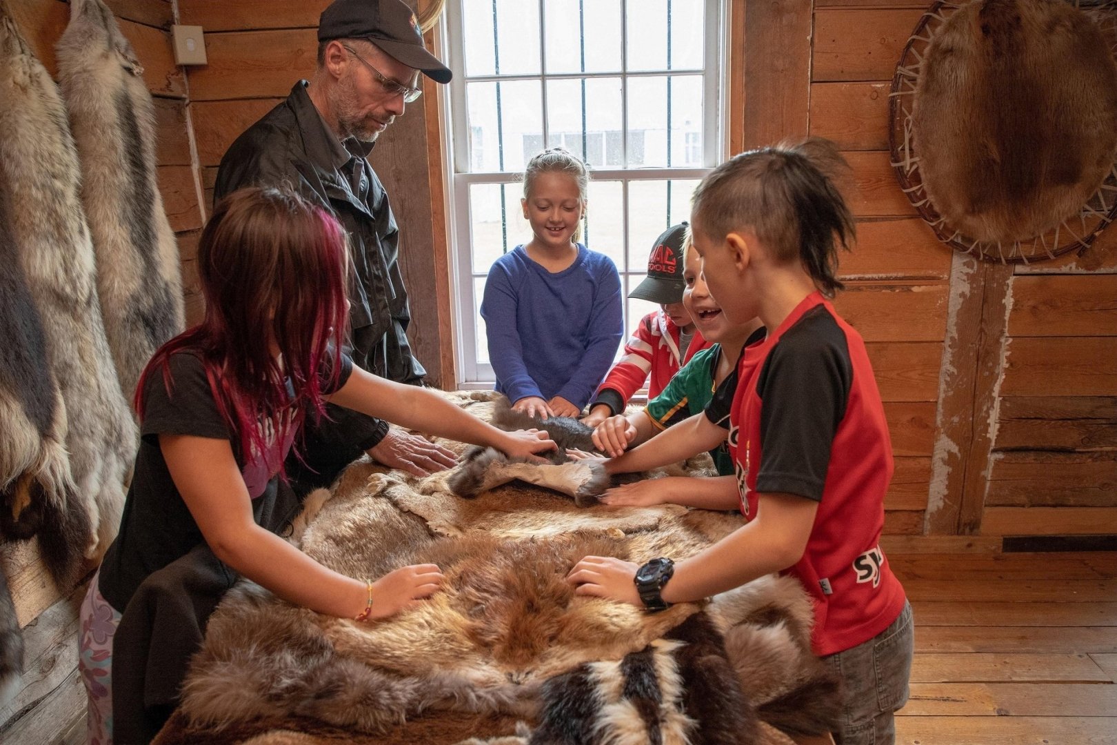 Kids playing with furs that would've been popular during the fur trade era, smiling and laughing while park interpreter smiles with them. 