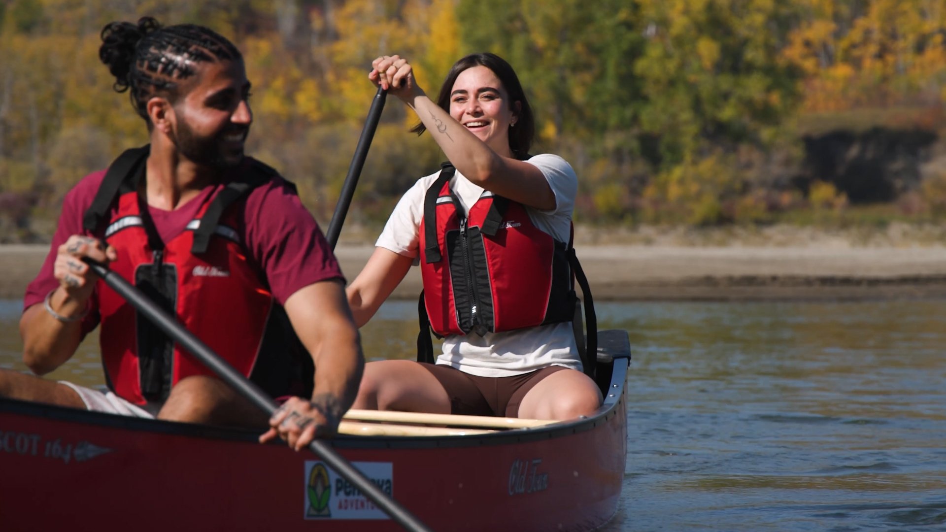 Man and women paddle in a canoe on still river smiling with fall leaves in the background