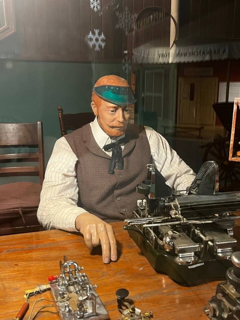 The image shows a mannequin portraying a telegrapher. He is sitting at a desk in front of a typewriter wearing a green visor and brown vest.