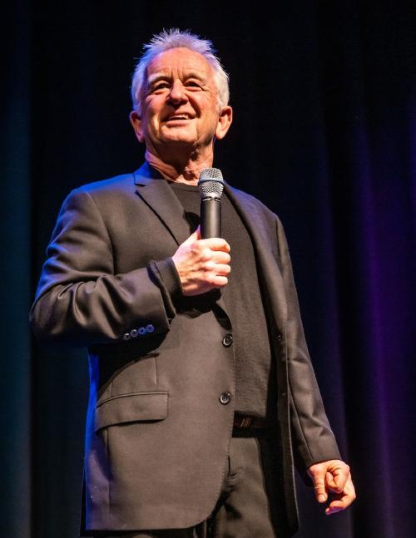Image of comedian Ron James looking out to the audience while holding a microphone to his face. 