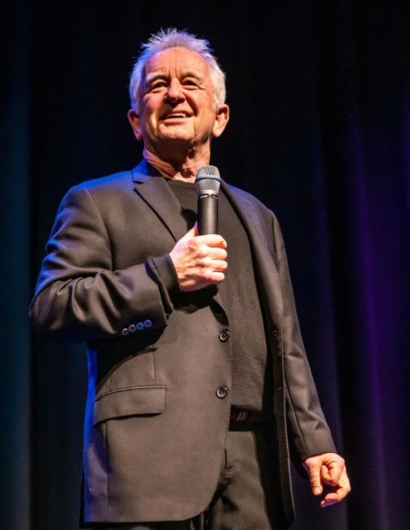 Image of comedian Ron James looking out to the audience while holding a microphone to his face. 