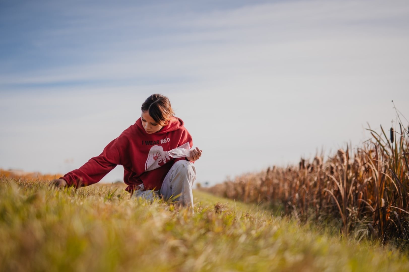 A young child wearing a red hoodie crouches in the grass looking curiously at the ground.