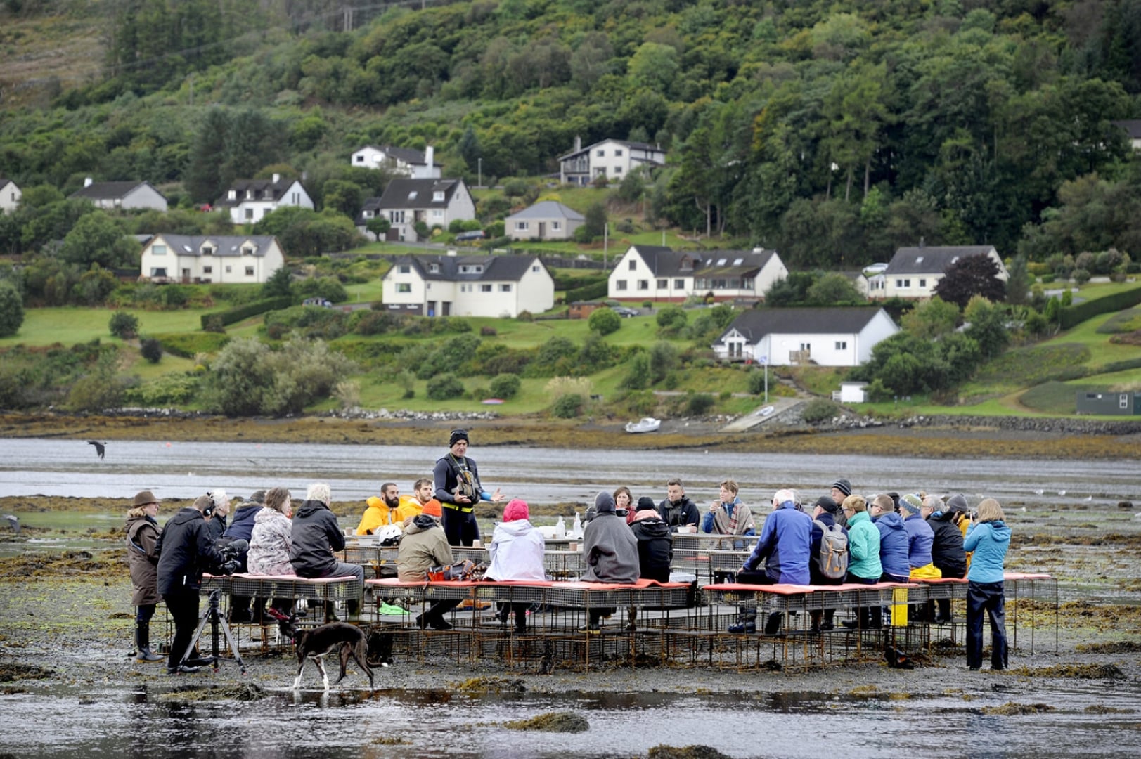 CLIMAVORE: On Tidal Zones. Public workshop by Rory MacPhee at low tide. A group of people gather around tables set on a tidal shoreline while a guide speaks at the center. Across the water, a small village and forested hillside create a scenic backdrop.