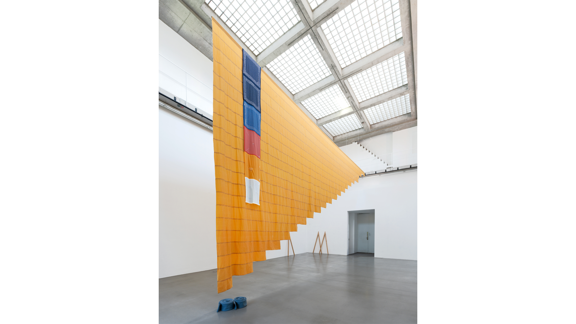 A large textile installation (Tanya Lukin Linklater, Crested, 2026) hangs from the ceiling of a bright gallery with a grid-patterned skylight. The suspended artwork features layered orange fabric with blue, red, and white elements, cascading diagonally across the open space.