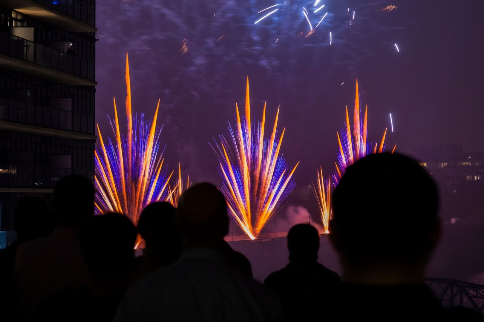 Silhouetted spectators watch a nighttime fireworks display, with bright blue and orange bursts lighting the sky. The colorful pyrotechnics rise above the water beside nearby buildings, creating a dramatic urban scene.