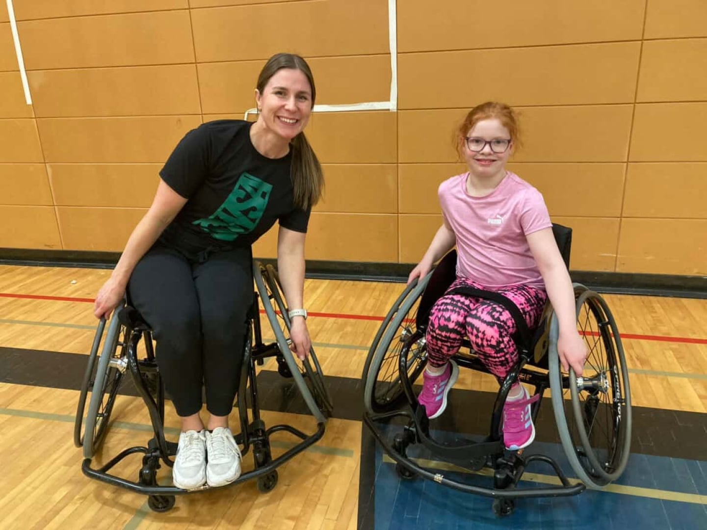 Female coach in a sport wheelchair beside a young girl in a sport chair. Both are smiling  looking at the camera and have their hands on the wheels.