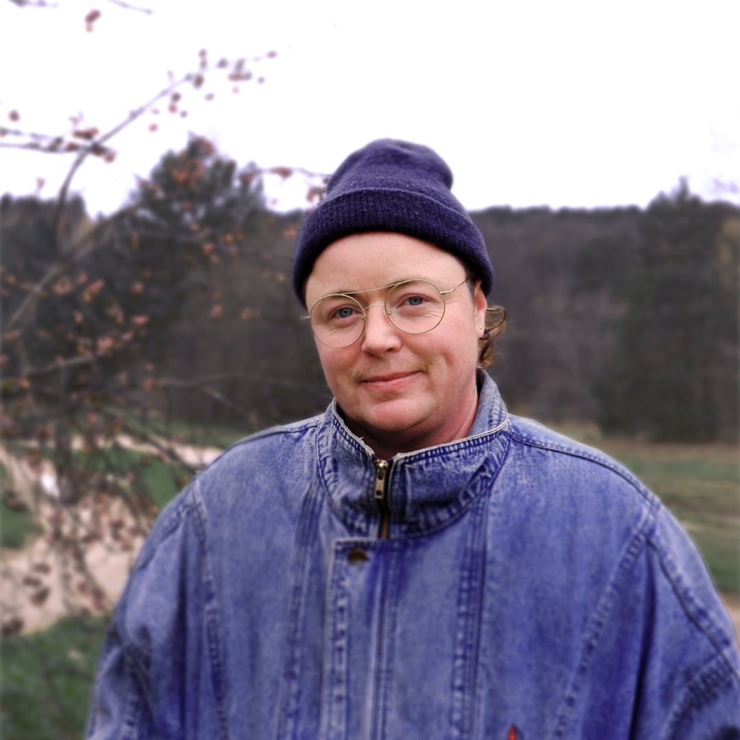Portrait of artist Lou Sheppard standing outdoors in front of a softly blurred landscape. Lou wears round glasses, a dark knit cap, and a blue denim jacket, with bare tree branches and a wooded background visible behind him.