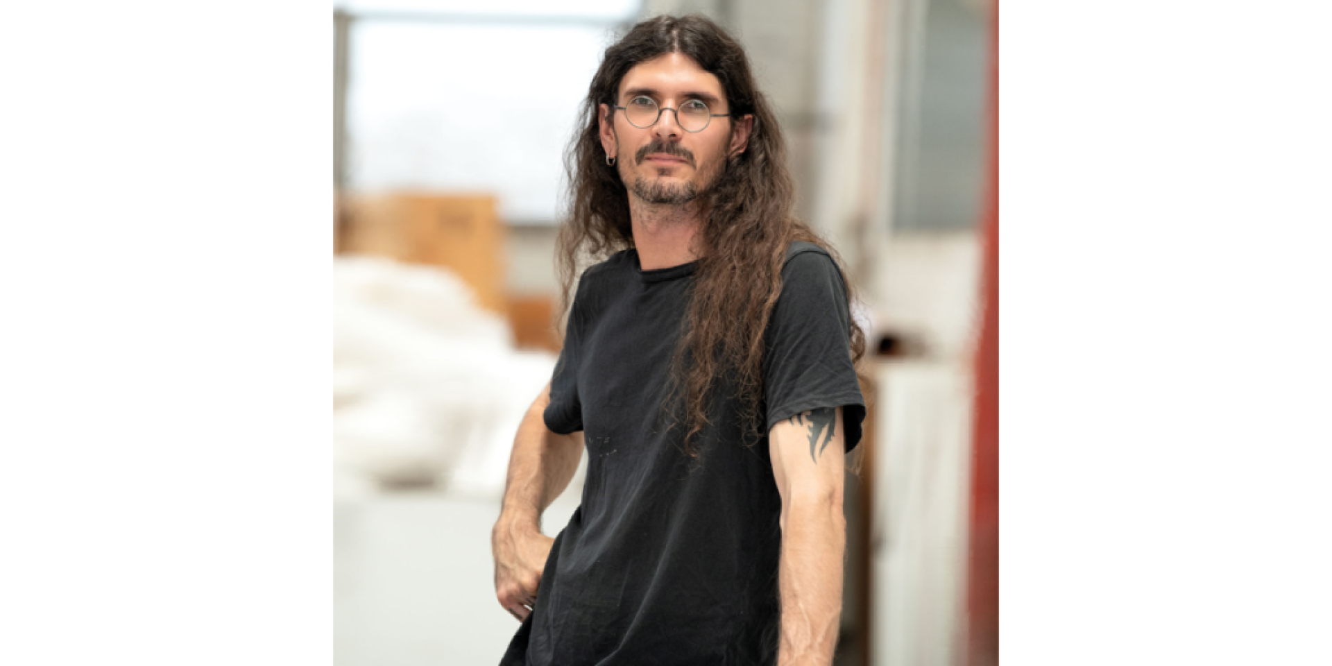 Portrait of artist, d harding, with long dark hair and glasses, wearing a black T-shirt and standing indoors. The person faces slightly to the side with one hand at their hip, while a softly blurred studio or workshop setting appears in the background.