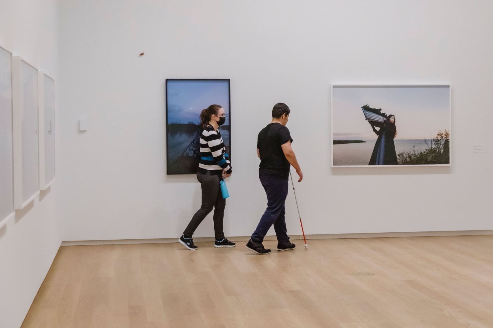 A participant uses a white cane to navigate an art gallery during a described tour while a guide walks alongside. Large framed photographs hang on the gallery walls as the group moves through the exhibition space.