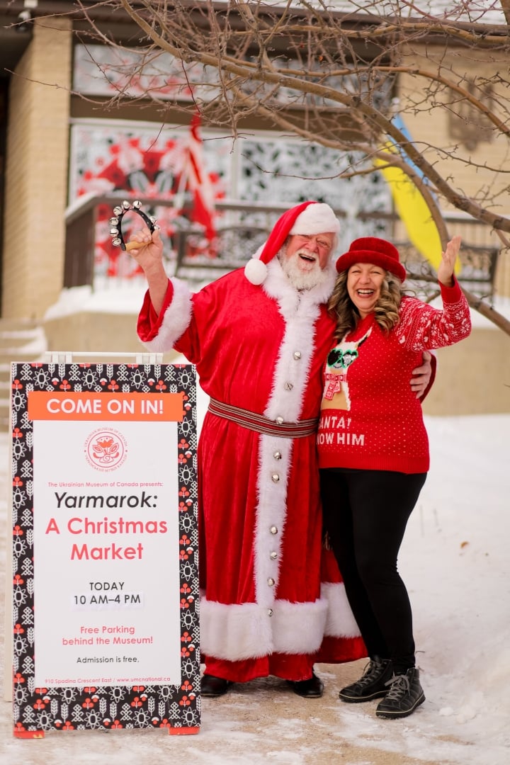 A photo of Santa and a volunteer outside of the museum next to the Yarmarok sign (sandwich board)