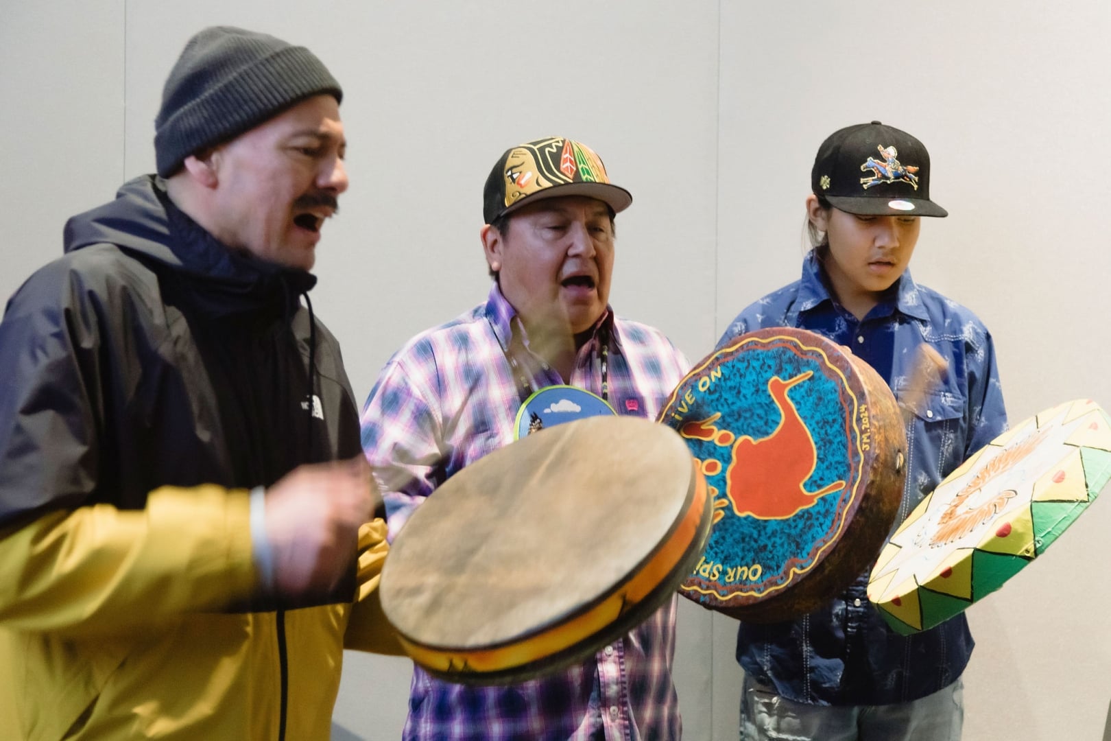Three people stand in a circle playing hand drums and singing during a cultural performance. The drums are brightly painted with colourful designs, and motion blur captures the rhythm and movement of the drumming against a simple indoor backdrop.