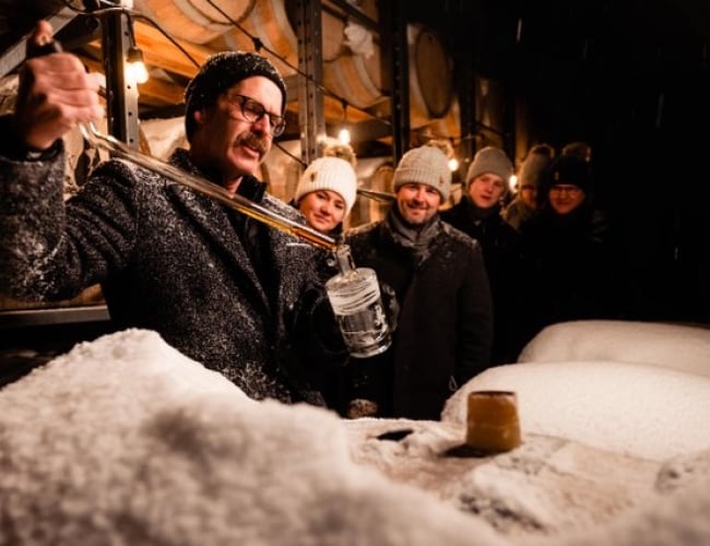 Man distilling whisky from a cask into a glass while onlookers marvel 